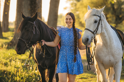 Beautiful woman with long hair walking with her horses in nature during sunset.