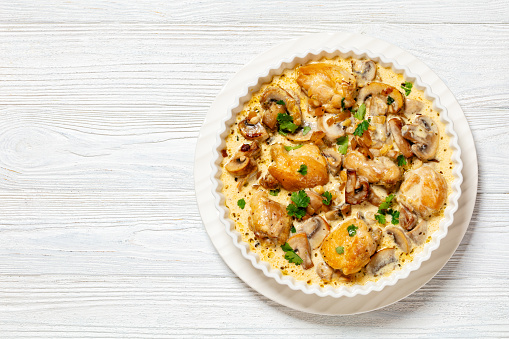 chicken thighs in a thick creamy mushroom garlic sauce with herbs and parmesan cheese in white baking dish on white wooden table, horizontal view from above, flat lay, free space