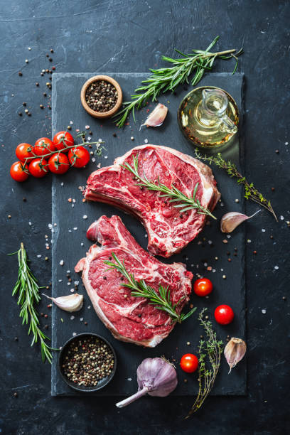 istock Two raw beef steaks on the bone on a black slate serving plate, decorated with various ingredients. Directly above view 2149218980