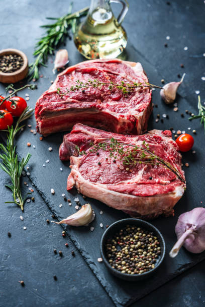 istock Two raw beef steaks on the bone with a thyme, and sea salt on the black slate serving plate. Decorated with a cherry tomatoes, garlic, pepper seasoning and decanter of cooking oil 2149219009