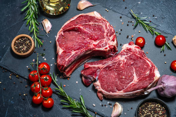 istock Two raw beef steaks on the bone on a black slate serving plate, decorated with various ingredients. 2149219084