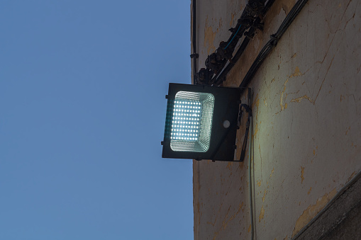 An LED lantern mounted on the street wall dimly illuminates the wall during dusk on dark blue sky background. The interval of time of day that is observed by a change in the degree of illumination.