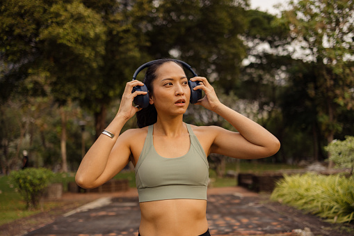 Portrait of pensive Thai sportswoman putting on headphones before sports training in the park.