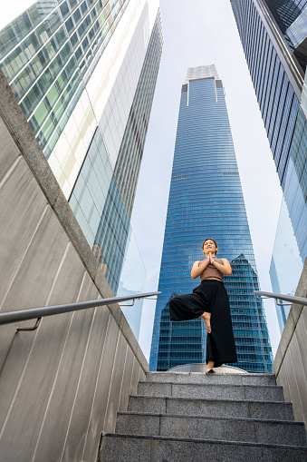 An active young Asian sportswoman practices mindfulness yoga outdoors in the morning, with a spectacular urban city skyline in the background. The concept embodies wellness, fitness, and a healthy lifestyle.
