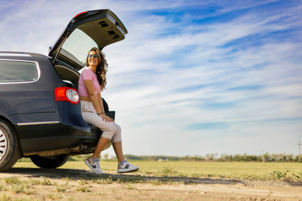 Young beautiful woman sitting in the trunk of her car. She is relaxed and happy while making a brake on her traveling through beautiful landscapes on sunny day