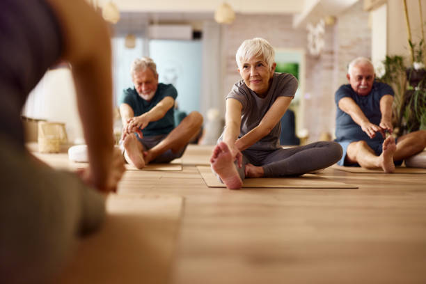 Active seniors stretching on exercise class in a health club. Group of mature people doing stretching exercises with their coach during a Yoga class in a studio. Focus is on senior woman. Copy space. yoga stock pictures, royalty-free photos & images