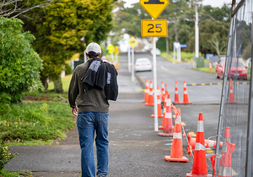 Man walking on a pedestrian footpath. Orange traffic cones and metal fences on the road. Road works in Auckland.