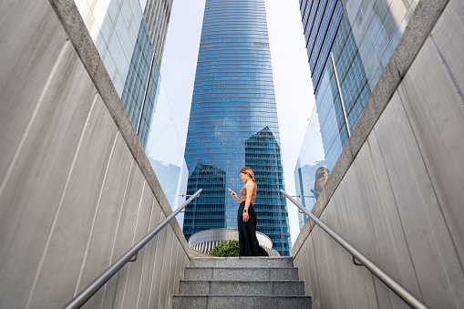 A young Asian woman seamlessly juggles urban life with the digital realm, as she converses over her smartphone amidst the towering buildings, bridging connections and networking effortlessly.