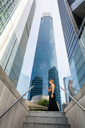 A young Asian woman seamlessly juggles urban life with the digital realm, as she converses over her smartphone amidst the towering buildings, bridging connections and networking effortlessly.