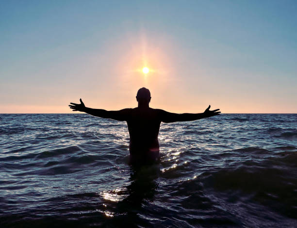 A man seen from behind, standing in the sea water and facing the sun, his arms outstreched in connection with the nature