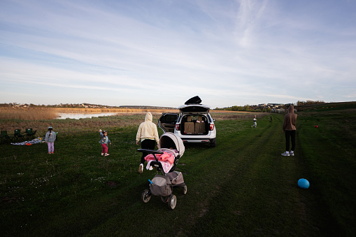 Serene family outing capturing three kids and adults near a parked car on a grassy field by a lake, enjoying the outdoors with toys and snacks.