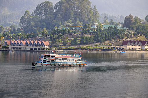 Lake Toba, North Sumatra, Indonesia - February 2nd 2024:  Ferry on the volcanic lake in front of a city with typical modern Batak architecture