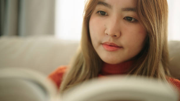 Asian young woman reading a book on sofa at home.