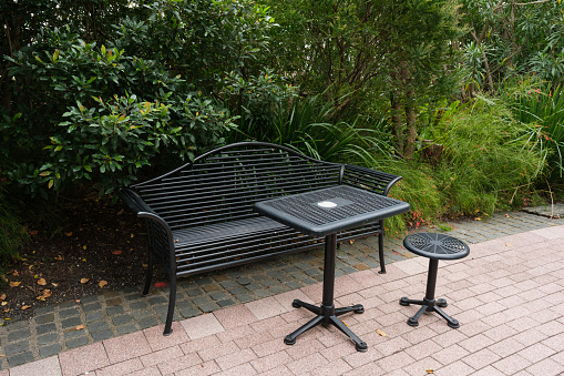 Black metal bench and tables on a paved walkway with trees and plants behind.