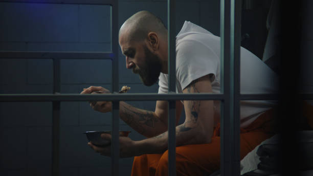 Male prisoner sits on bed in prison cell, eats disgusting food Male prisoner in orange uniform sits on bed in prison cell, eats disgusting prison food from iron bowl. Inmate serves imprisonment term for crime in jail. Detention center or correctional facility. prison meal stock pictures, royalty-free photos & images