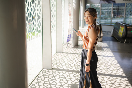 A young Asian woman seamlessly juggles urban life with the digital realm as she converses over her smartphone amidst the pedestrian walkway, effortlessly bridging connections and networking while looking at the camera.