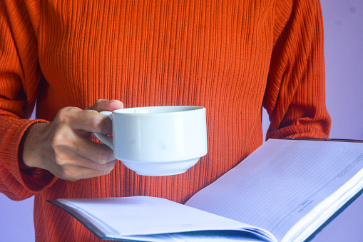 Portrait of a woman wearing an orange t-shirt doing her morning activities by drinking a cup of black coffee and reading a book