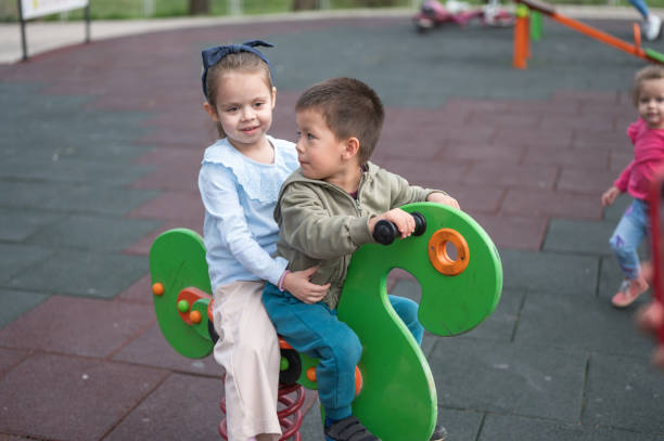 A boy and girl smiling, sharing a spring rider at the playground with motion blur in the background. Multiracial group of toddlers playing in the park