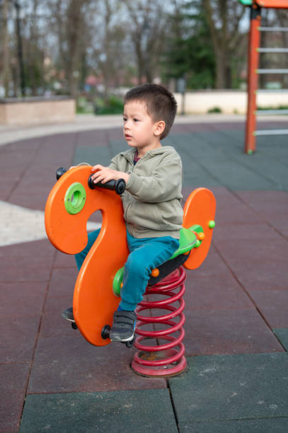 A 3 year old multiracial boy concentrating while sitting on an orange and green spring rider alone, with playground structures behind