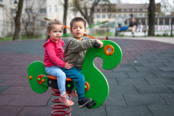 A pair of young children sharing a green spring rider with a look of companionship, in a playground. Two and three year old girl and boy playing in the park. Capture the essence of childhood friendship as two young children enjoy a playful moment together on a green spring rider in a vibrant playground