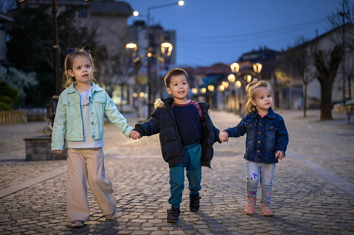 Night scene of two girls and one boy holding hands and joyfully walking on a cobblestone street. Two girls and one boy hold hands and run joyfully on a cobblestone street at night, their laughter echoing through the illuminated surroundings. Toddlers having a fun time out