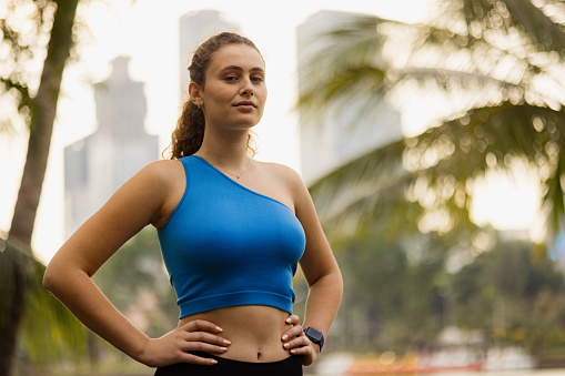 Young sportswoman standing with arms on her hips during a break in the park and looking at camera.