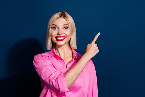 Photo of cheerful woman with bob hairstyle red lips dressed pink shirt indicating at discount empty space isolated on dark blue background.