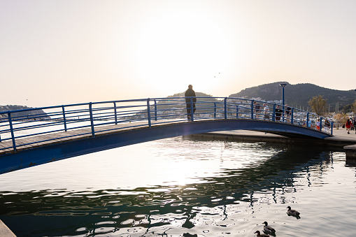 Puerto Andratx, Spain -April 14, 2024: Afternoon in Andratx harbor with a bridge and walking people against the setting sun with two ducks swimming in the water. The harbor Puerto Andratx, Mallorca, is renowned for its affluent international visitors, whether tourists or residents, especially during sunset and for its pub nightlife. Regularly, there is live music or small attractions. The restaurants and bars are almost fully booked every evening. The port of Andratx is considered the Saint-Tropez of Mallorca.