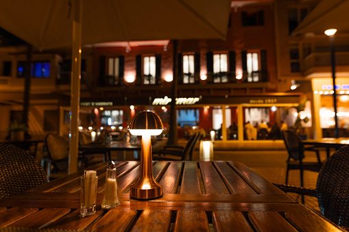 Puerto Andratx, Spain -April 14, 2024: Beautiful close up of a restaurant table at night at Andratx's port with unfocused guests sitting outdoors in the restaurant. The harbor Puerto Andratx, Mallorca, is renowned for its affluent international visitors, whether tourists or residents, especially during sunset and for its pub nightlife. Regularly, there is live music or small attractions. The restaurants and bars are almost fully booked every evening. The port of Andratx is considered the Saint-Tropez of Mallorca.