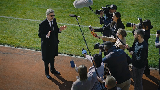 SEO or director of football team answering press questions and giving interview near soccer field before important match. Private investor or businessman crowded by news journalists. Press conference.
