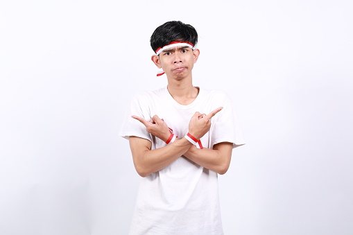 Confused Asian young boy with red white ribbon for celebrating Indonesia independence day standing while pointing right and left side