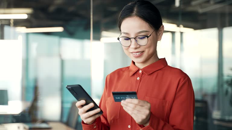 Happy young asian female employee doing online shopping using credit card and mobile phone while standing in business office.