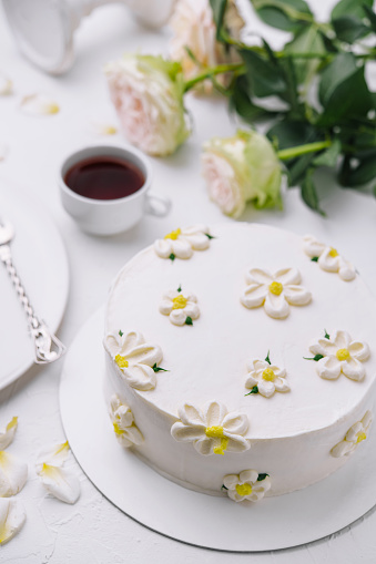 White frosted cake adorned with flowers, alongside tea and roses, on a chic table setting