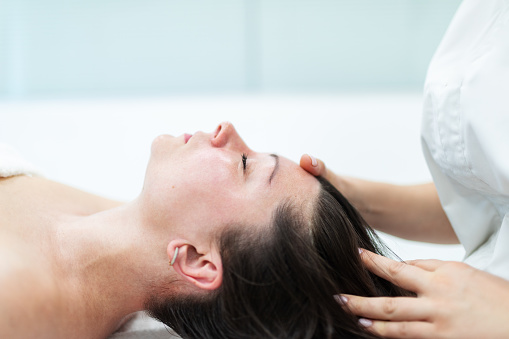 Cosmetologist making facial massage to mid adult woman, mid adult woman lying down on massage table relaxed and happy