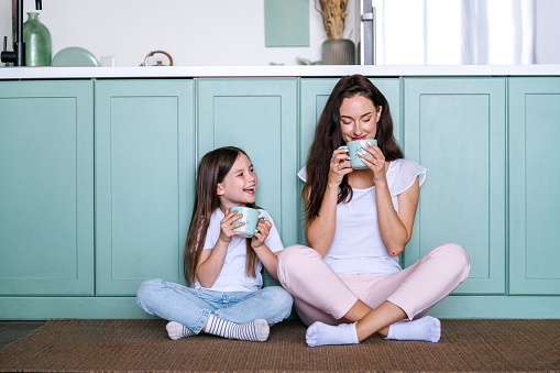 Dreamy mother sitting on kitchen floor together with daughter, drinking coffee, enjoying morning beverage and spending weekend in cozy apartment, resting at home. Smiling kid looking at mom