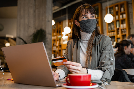 Portrait of a young woman wearing a face mask and making a credit card purchase while using her laptop