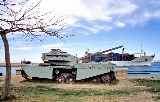 Old rusty armor tank alongside the Suez canal in Egypt with passing container ships at the background