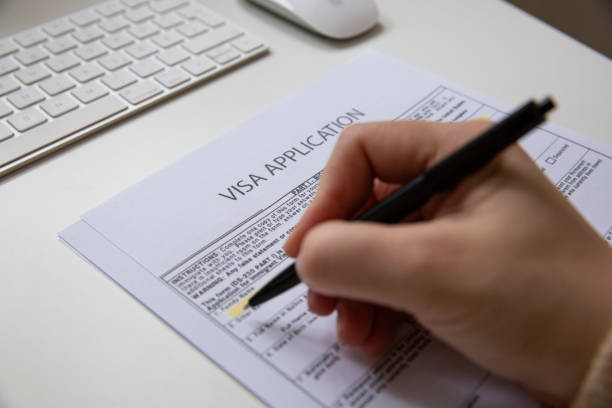 istock Close up of a female hand filling out a visa application document, white table with computer keyboard and a mouse in background 2149247772