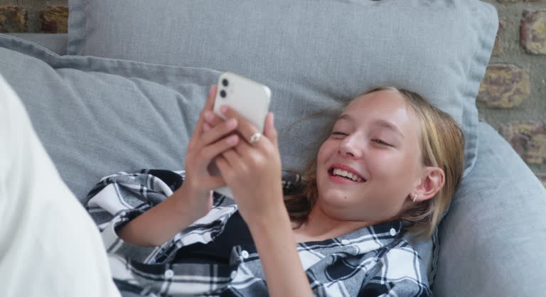 Happy teenage girl laughing while using smartphone on sofa at home