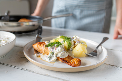 Fresh cooked healthy homemade meal with salmon fillet, boiled potatoes and light yogurt herb sauce with woman in the background. Served on kitchen counter.