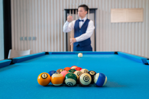 A sharply dressed man in a vest and bow tie focuses intently while lining up a shot on the blue felt of a billiard table.