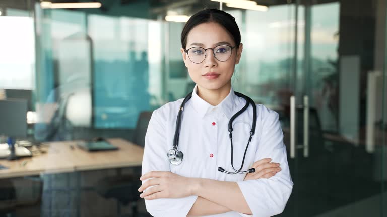 Portrait of young serous asian female doctor in white coat and glasses looking at camera in modern hospital clinic.