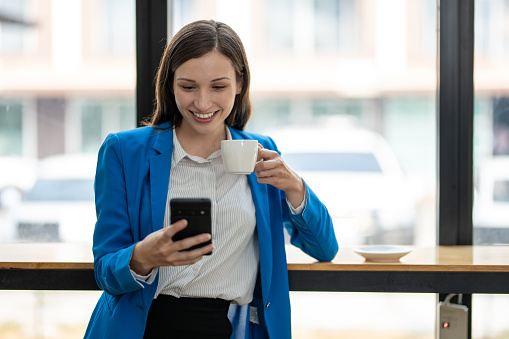 Happy professional female in a blue suit enjoying a coffee while using her smartphone in an office setting.