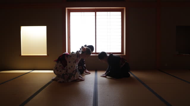 Female tourists in kimono learning manners of greetings from a senior instructor in dark Japanese tatami room