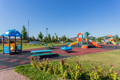 A children's playground with bright playsets and benches against a backdrop of greenery and blue sky.