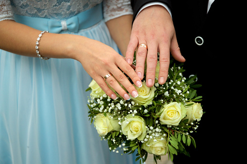 The hands of the bride and groom with wedding rings against a wedding bouquet of white roses.