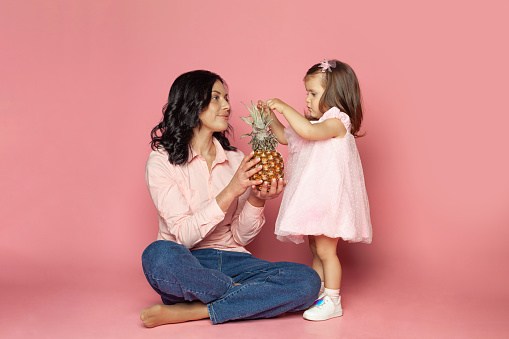 Young brunette woman and baby girl holding fruit on pink background