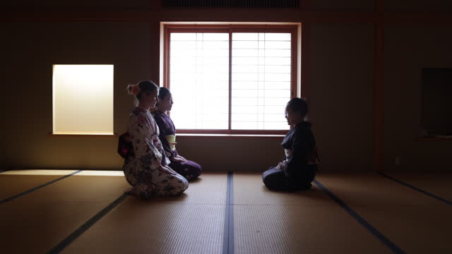 Female tourists in kimono learning manners of greetings from a senior instructor in dark Japanese tatami room