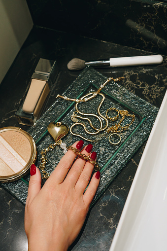Gold necklaces, rings and bracelets next to liquid foundation and shimmering highlighter on the glass plate on dark marble counter. Personal luxury and style concept. Elegant woman's hand take a bracelet from the plate.