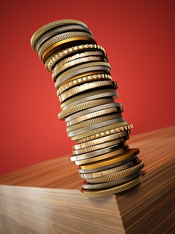 A tall, unstable tower of mixed coins balances on the edge of a polished wooden surface, with a vivid red backdrop highlighting the metallic colors and textures. High Investment Risk Concept.